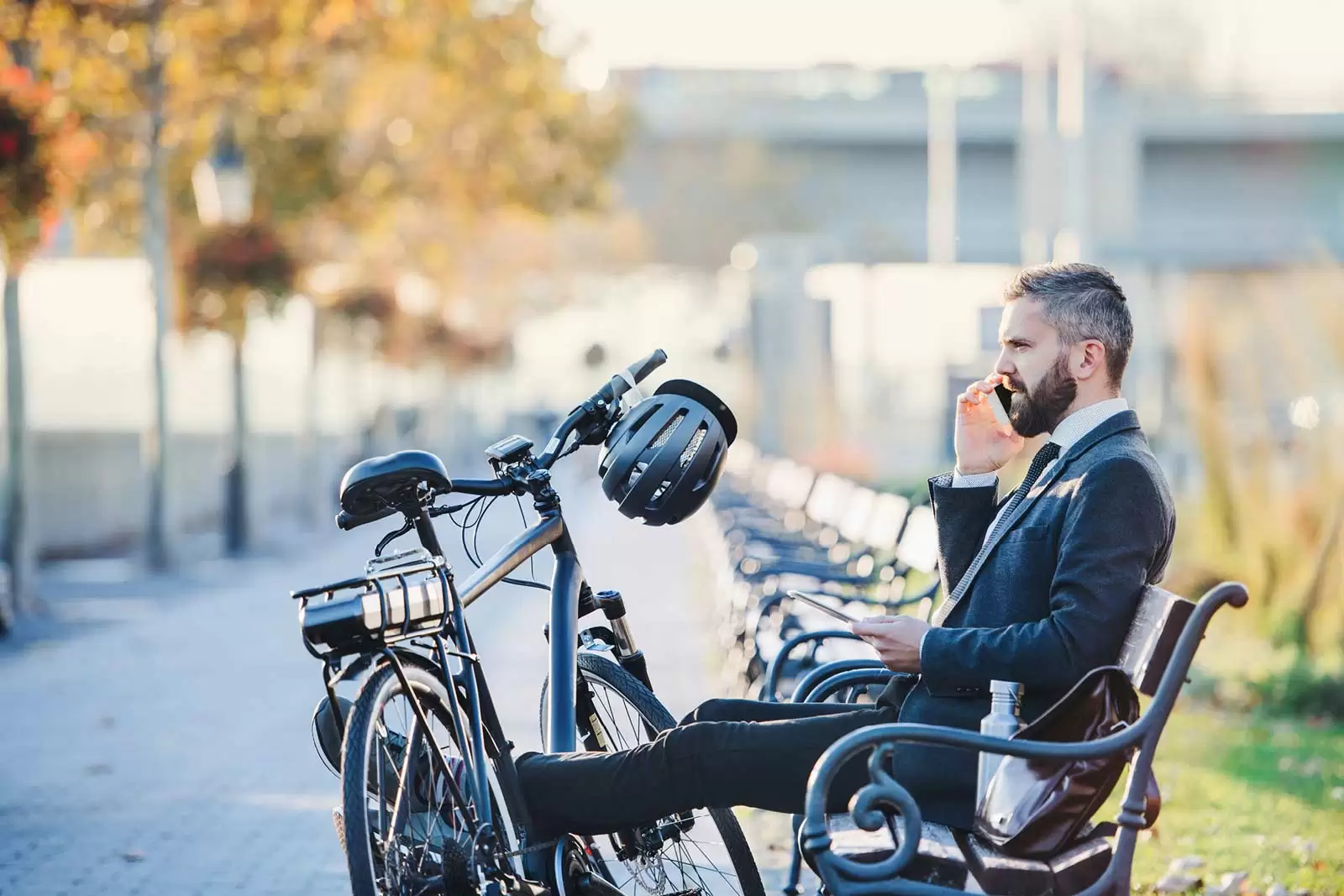 Geschäftsmann sitzt hinter seinem geleasten E-Bike auf einer Bank und telefoniert.